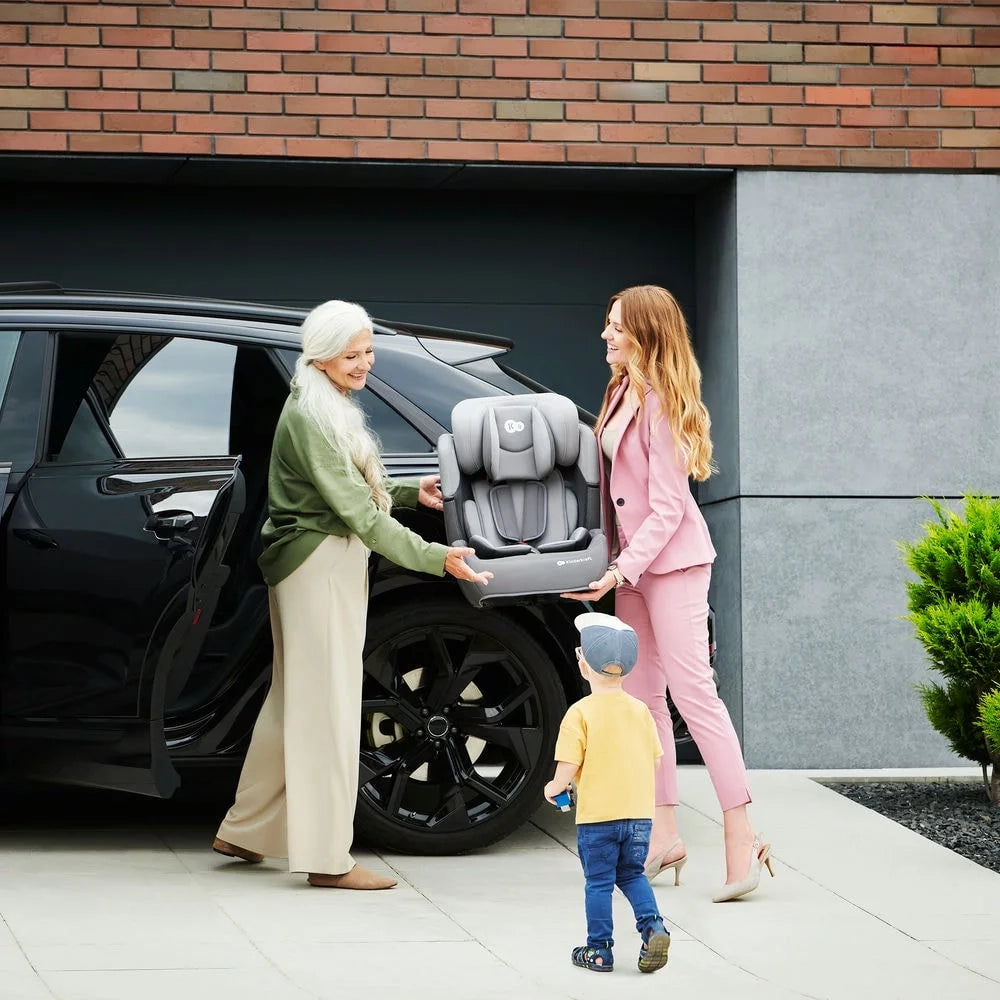Grandmother and mother exchanging a Kinderkraft car seat next to their car, with a toddler watching.