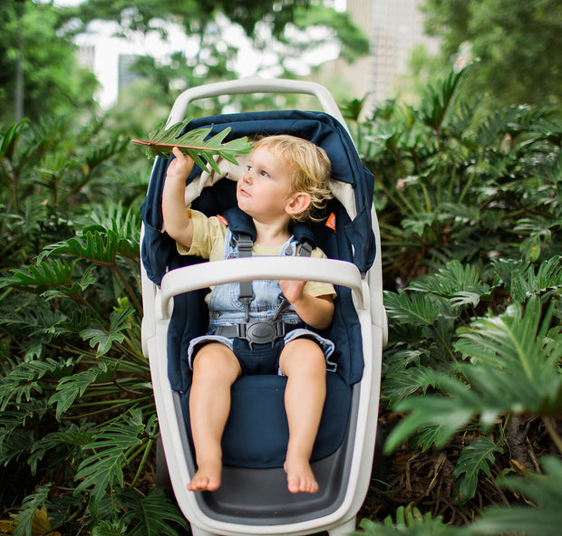 Toddler in Greentom Reversible stroller touching leaf, surrounded by lush greenery.