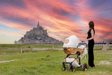 Woman standing with a Junama Diamond stroller in front of a picturesque castle at sunset, with vibrant skies and grassy foreground.