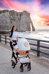 Woman pushing a stylish Junama Diamond stroller along a seaside boardwalk at sunset, with dramatic cliffs in the background.
