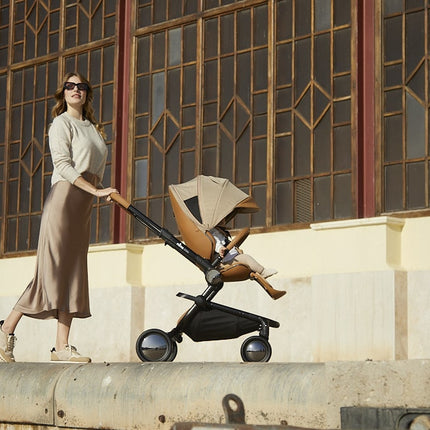 Woman pushing a Mima Creo Stroller Mocha with a child in it, enjoying a walk in the park.