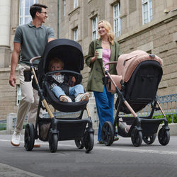 Parents strolling with Kinderkraft MOOV 2 strollers, one black and one pink, on a city street with their children.