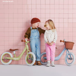 Two children with Kinderkraft RAPID balance bikes, green and blue, standing against a pink tiled wall.