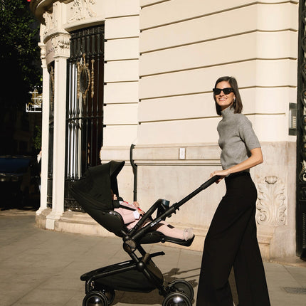 Woman in grey adjusting Mima Creo Stroller Black with toddler in pink outside a historic building on a sunny day.