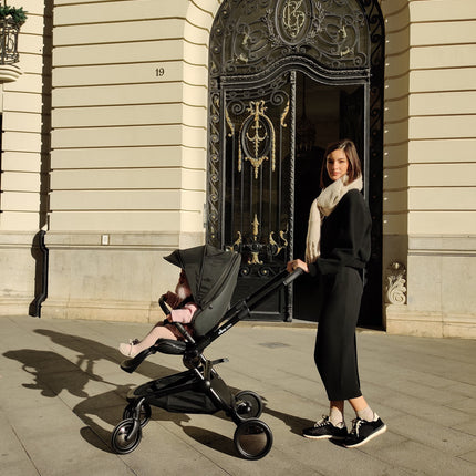 Woman in black with toddler in pink outfit using Mima Creo Stroller Black on a city stroll outside a historic building.