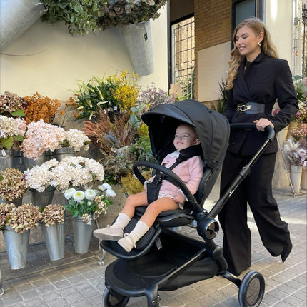 Woman in black with smiling toddler in pink outfit using Mima Creo Stroller Black near a flower shop.