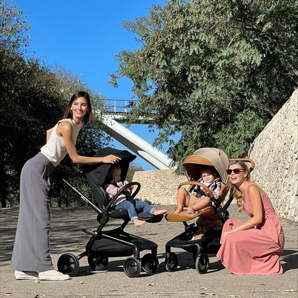 Two women with toddlers using Mima Creo Stroller Black during a park walk, one in pink hoodie, one in straw hat