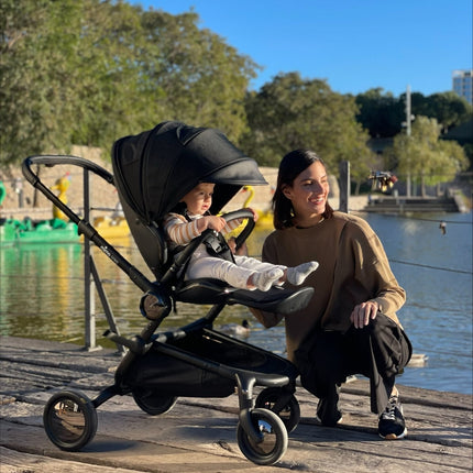 Woman in beige with toddler in striped shirt using Mima Creo Stroller Black by the riverside, enjoying the view.