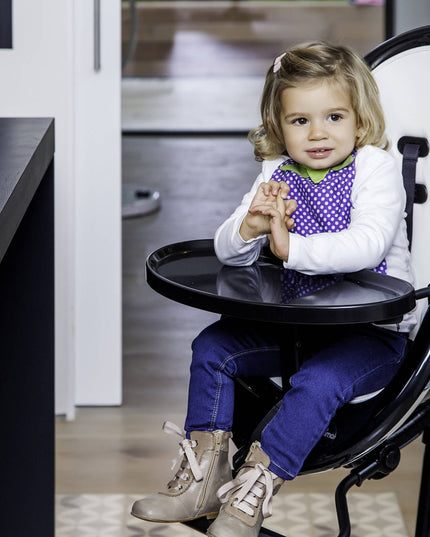 Child enjoying a snack in a Mima Moon High Chair with a white cup and cookies on the table.