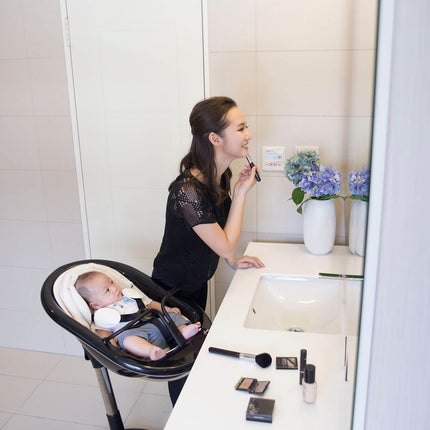 Mother applying makeup with baby in a Mima Moon High Chair in the bathroom.