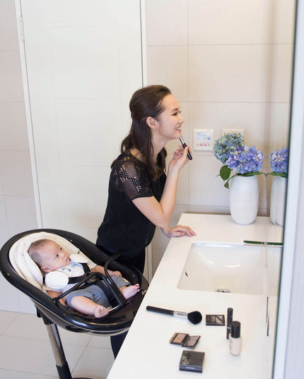 Mother applying makeup with baby in a Mima Moon High Chair in the bathroom.