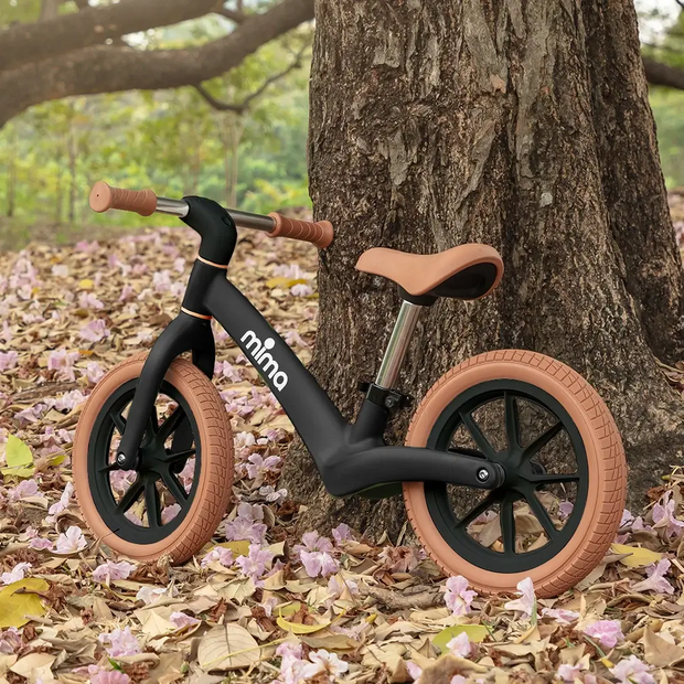 Mima ZOOM Lite Balance Bike in black with brown accents, placed by a tree