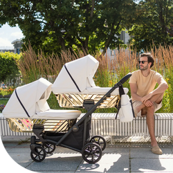 Man sitting beside a white double stroller with gold accents, set in an urban park, highlighting a contemporary and luxurious stroller.