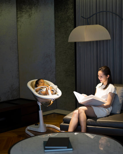 Woman reading a book next to a Mima Moon High Chair with baby in a cozy living room.